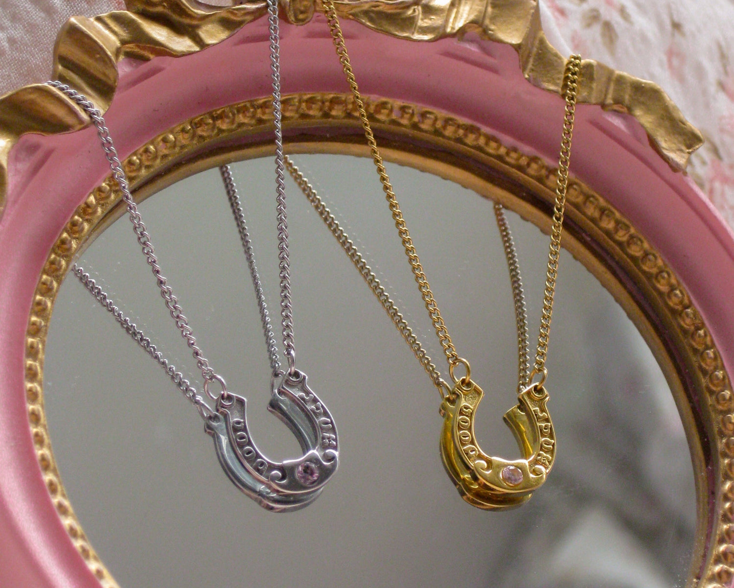 Pink mirror with gold frame displaying two necklaces, one silver and one gold, on a floral background.